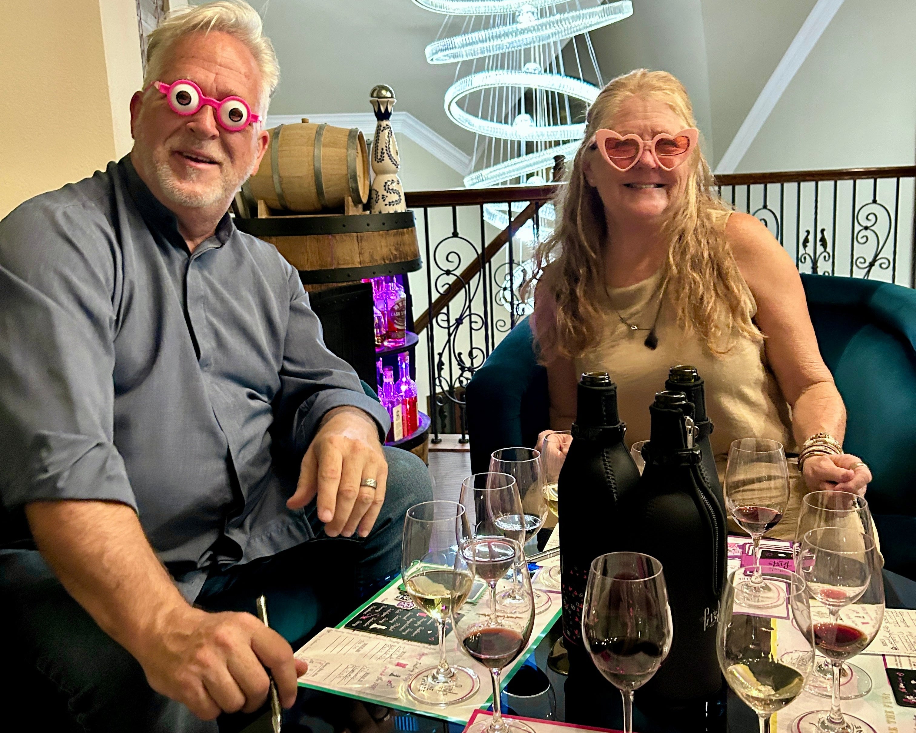 Two people sitting at a table with wine bottles and glasses, wearing novelty sunglasses and playing FMK.