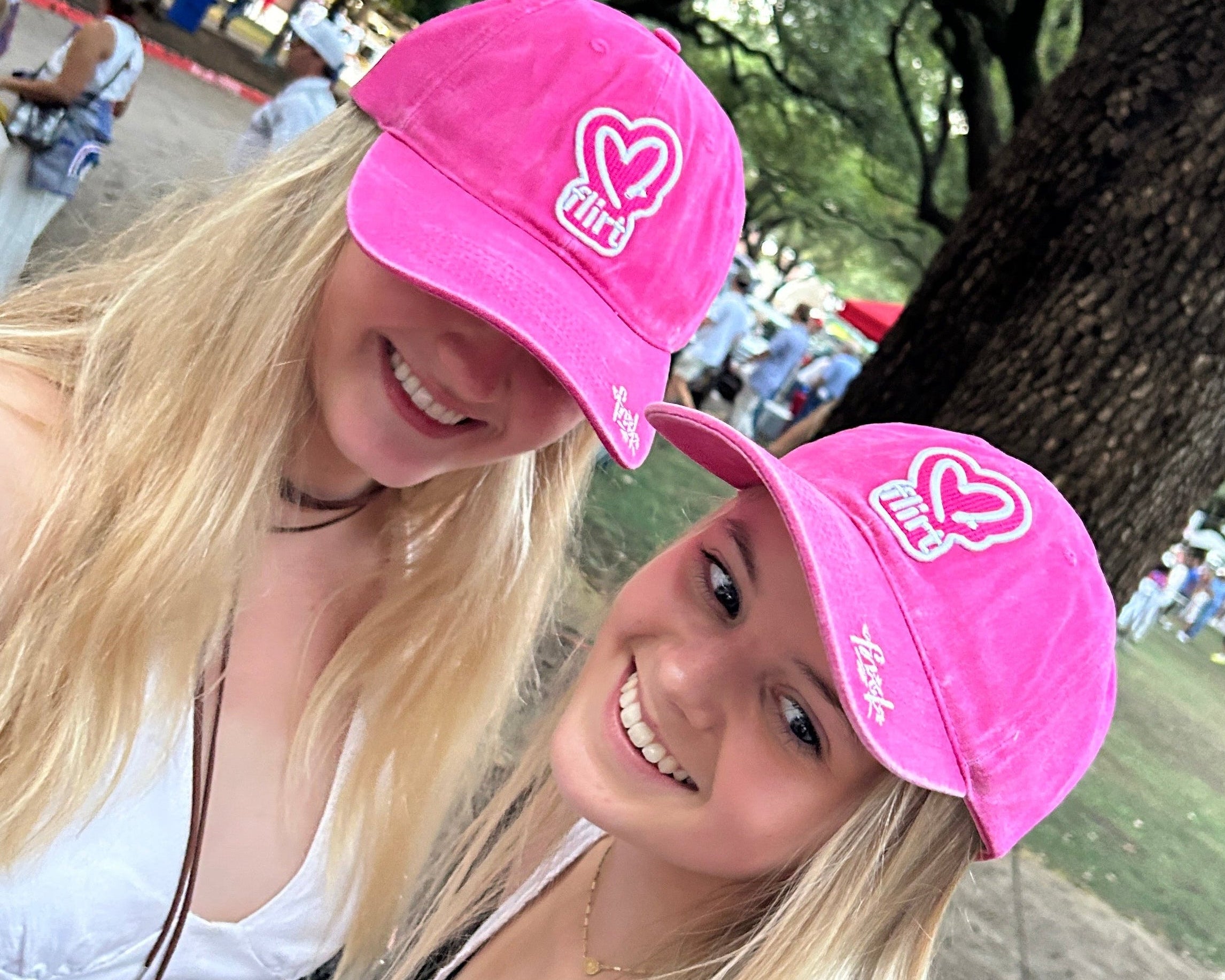 Two women wearing pink hats with a heart design, standing outdoors near trees and a water feature.